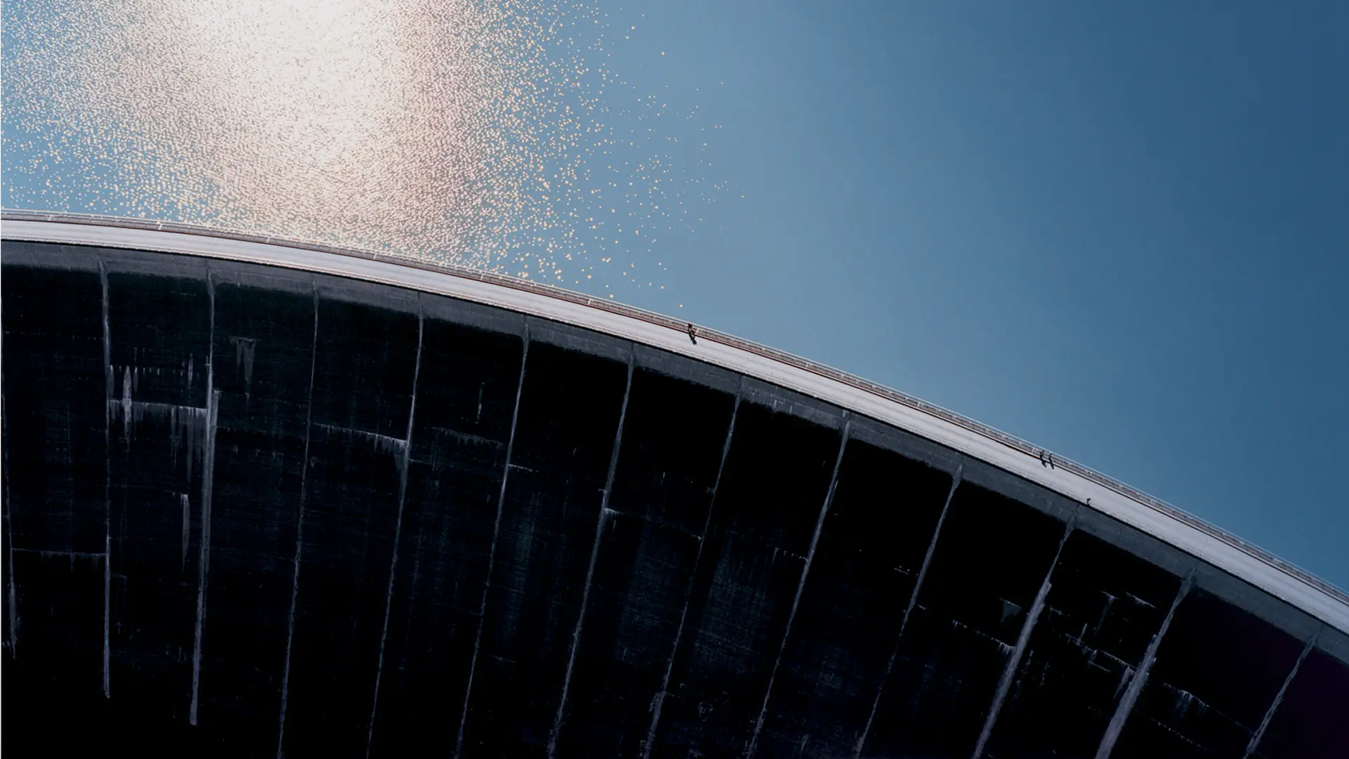 Aerial view of a large dam; the sun reflects on the reservoir, and pedestrians are walking on the dam.
