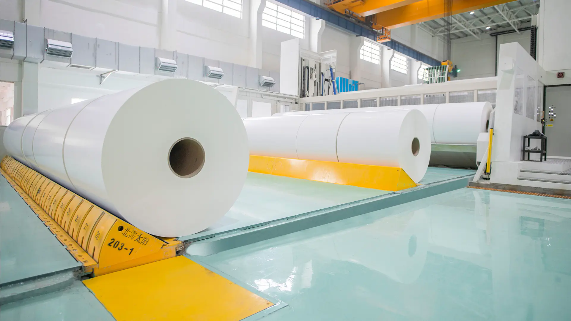 Interior view of a modern paper mill; large rolls of white paper lie in the hall, with cranes and technical equipment visible on the ceiling and walls.